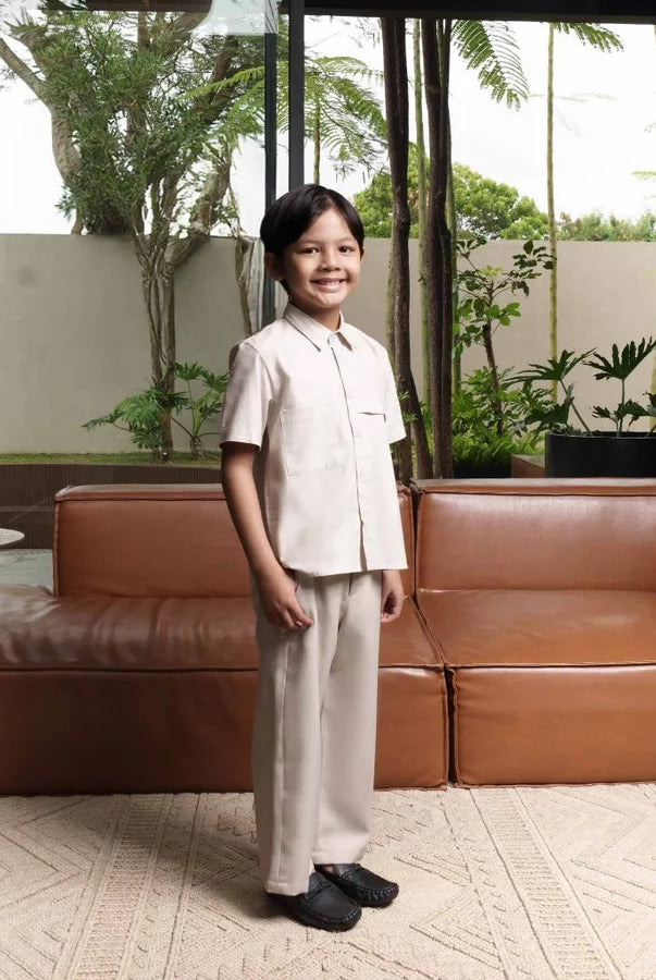 Child standing in front of a brown leather sofa wearing beige shirt by Jenna & Kaia in the background