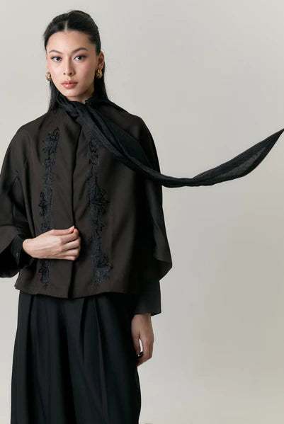 Woman wearing a black outfit with embroidery by Studio Tui and a long black scarf against a plain background