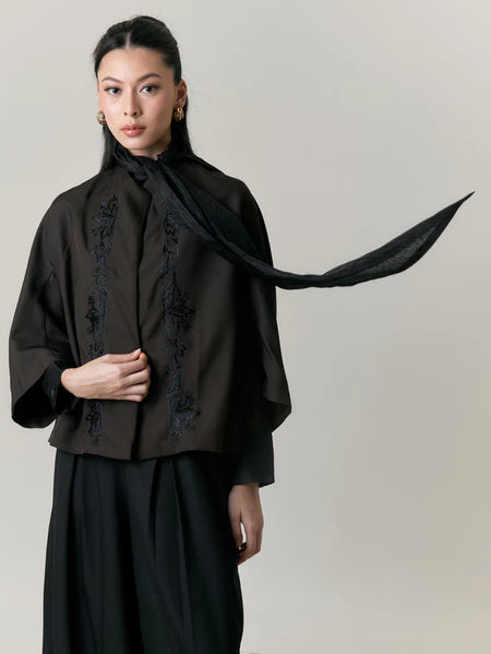 Woman wearing a black outfit with embroidery by Studio Tui and a long black scarf against a plain background