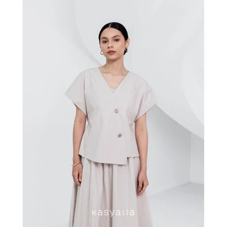 A woman wearing a beige, asymmetrical button-down blouse with short sleeves and a round neckline by Kasyalia.