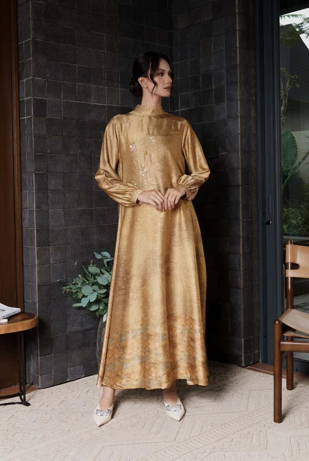 Woman in a gold dress by Jenna & Kaia standing in a modern indoor setting with a dark wall and wooden furniture.