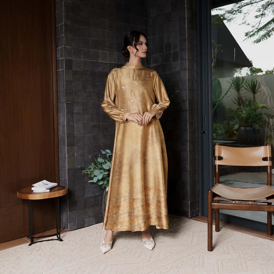 Woman in a gold dress by Jenna & Kaia standing in a modern indoor setting with a dark wall and wooden furniture.
