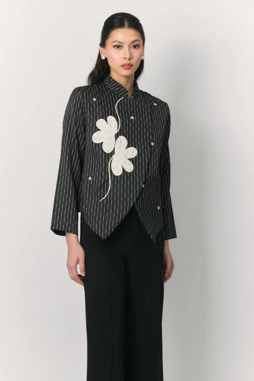 A model wearing a black and white vertically striped top by Studio Tui with a large white embroidered flower on the chest, paired with black trousers, against a plain light gray background.