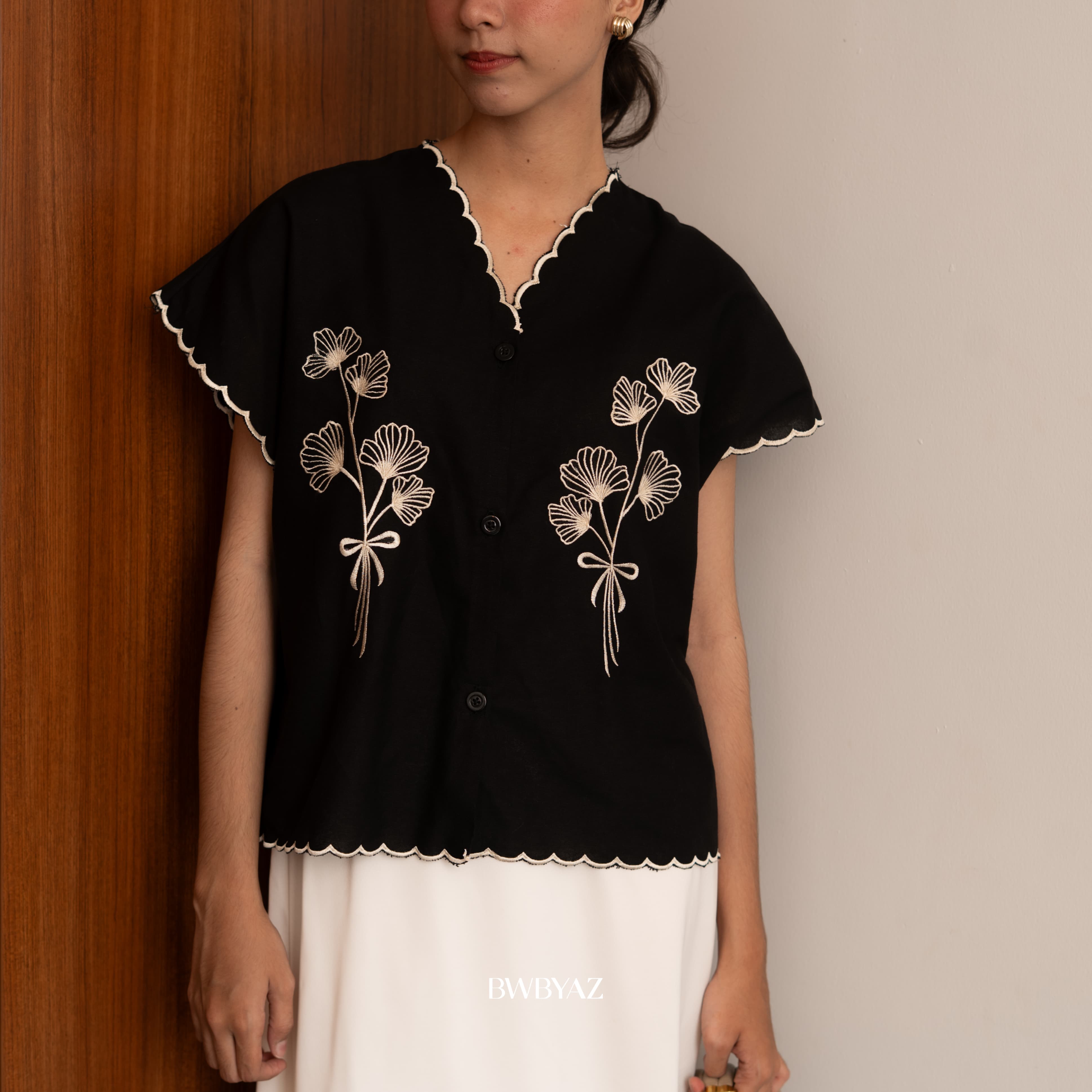 A person wearing a black Alaia Embroidery Tie Vest with white floral embroidery and a scalloped edge from BWBYAZ, paired with a white skirt.