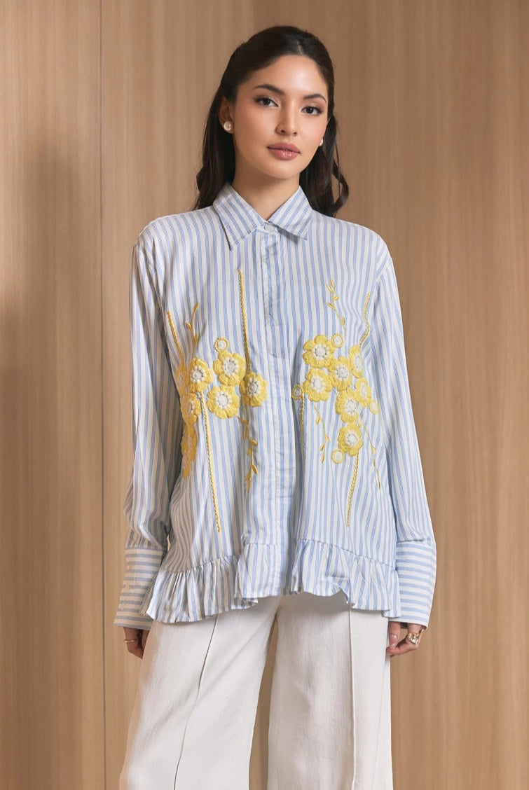 A woman models a light blue and white vertically striped long-sleeve shirt by Studio Tui with yellow floral embroidery on the front, paired with white trousers, set against a warm wood-paneled background.