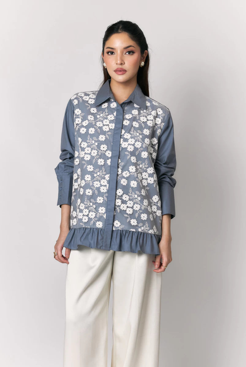 A woman models a slate blue button-up shirt by Studio Tui with a white floral embroidered pattern over the front panel, paired with cream wide-leg trousers, against a light gray studio background.