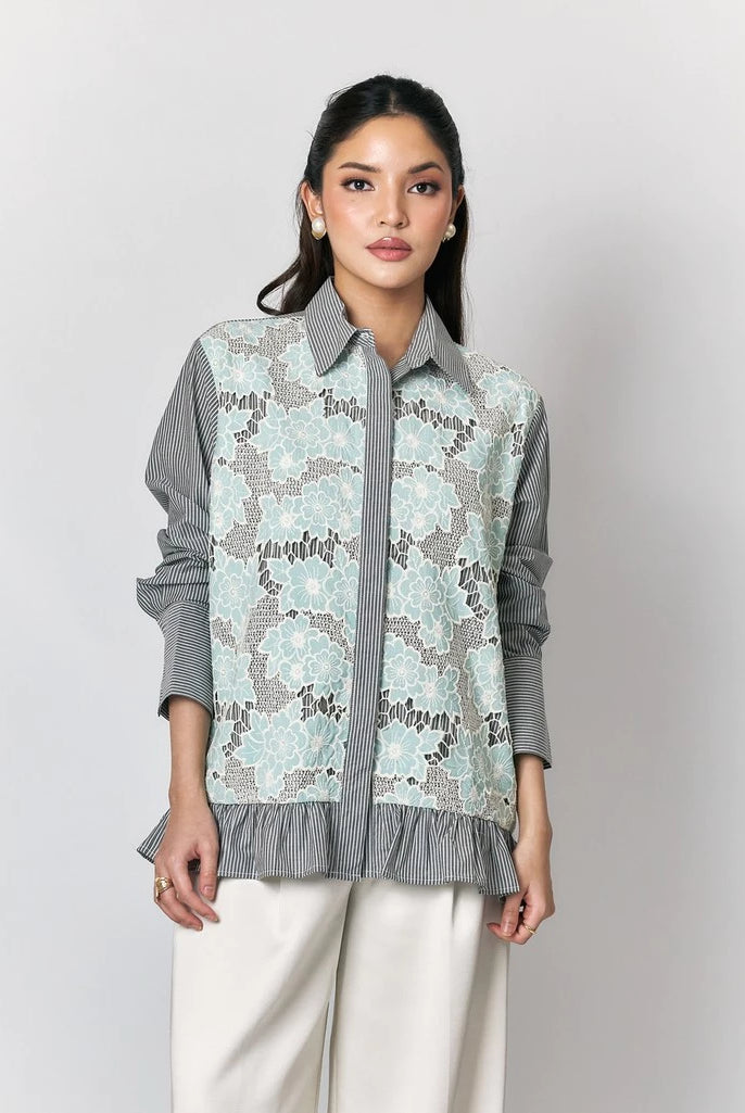 A woman models a long-sleeve button-up shirt by Studio Tui with a light blue and gray floral lace overlay on a gray striped base, paired with white trousers, against a neutral gray background.