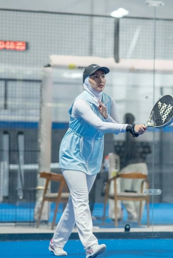 Woman wearing a modest sporty outfit featuring a light blue sleeveless vest by Abame with zipper details, a light-colored long-sleeve inner top, white flare pants, and white sneakers at an indoor padel court.