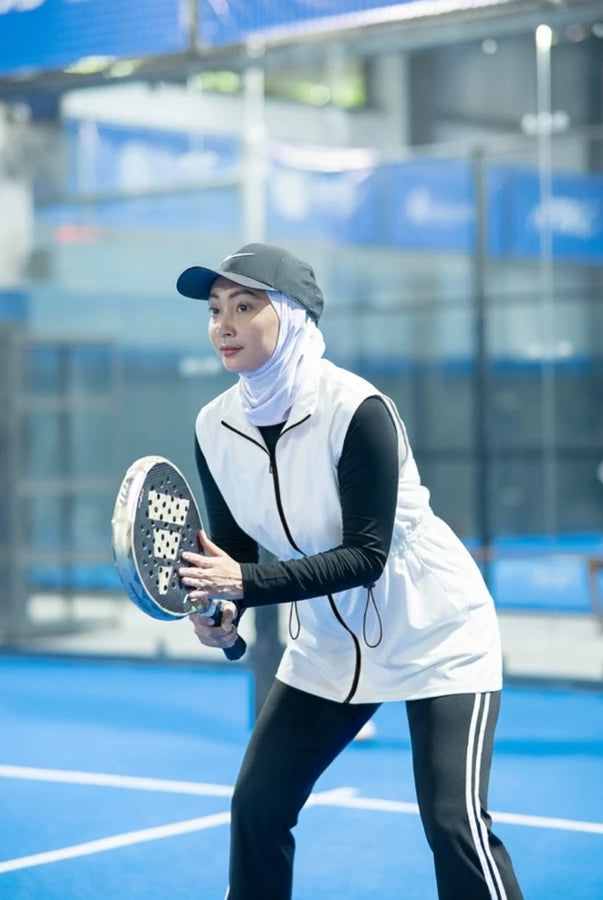 Woman wearing a modest sporty outfit featuring a broken white sleeveless vest by Abame with zipper details, a light-colored long-sleeve inner top, and black flare pants at an indoor padel court.