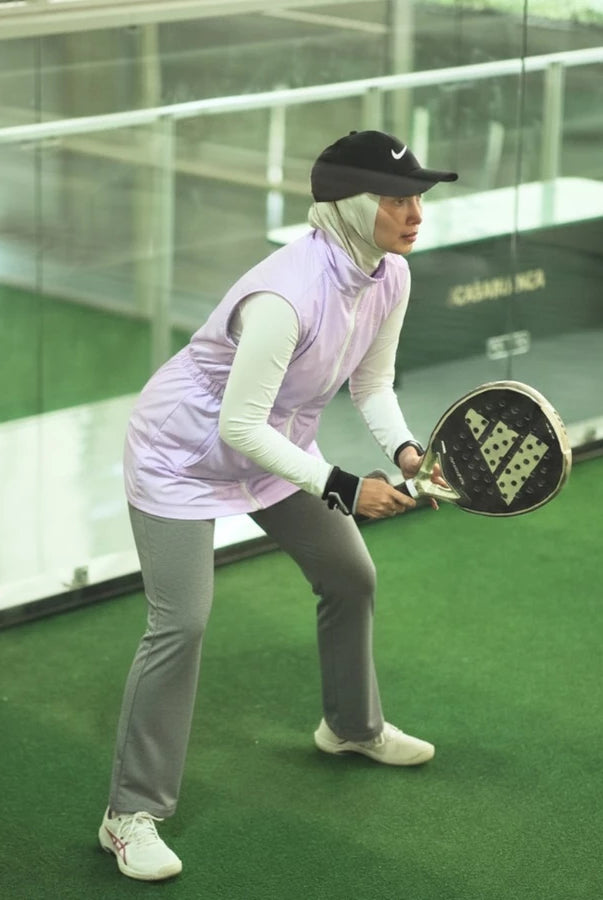 Woman wearing a modest sporty outfit featuring a lilac-colored sleeveless vest by Abame with zipper details, a light-colored long-sleeve inner top, light grey flare pants, and white sneakers at an indoor padel court.