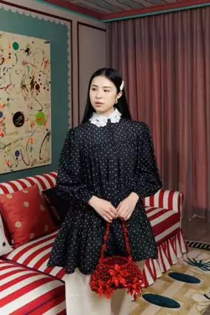 Woman in a black polka dot blouse by Haidee Orlin holding a red bag in a room with colorful walls and a striped couch.
