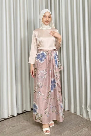 Woman wearing a floral skirt by Kartinis Label and beige outfit standing against a plain curtain background