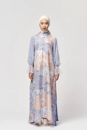 A woman standing in a studio setting, wearing a long, floral and geometric patterned dress by Kartinis Label with a denim-like texture.