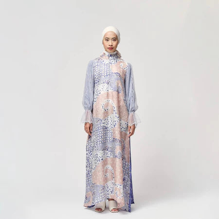 A woman standing in a studio setting, wearing a long, floral and geometric patterned dress by Kartinis Label with a denim-like texture.