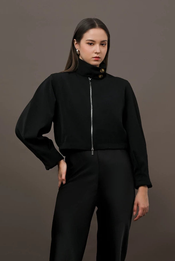 A model wearing a black cropped jacket by Kasa Heritage with a high mock neck, silver front zipper, and decorative gold buttons near the collar, paired with black trousers, set against a solid dark brown studio background.