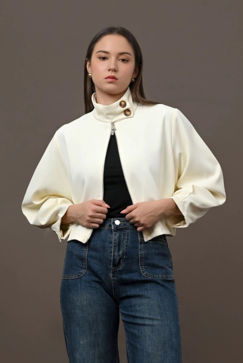 A woman modeling a cream-colored cropped jacket by Kasa Heritage with a high mock neck and gold-tone buttons, worn over a black top and blue jeans, against a solid dark gray studio background.