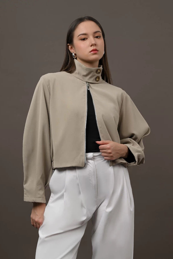 A woman modeling a cropped, high-neck beige jacket by Kasa Heritage with a front zipper, worn over a black top and white trousers, set against a solid dark gray studio background.