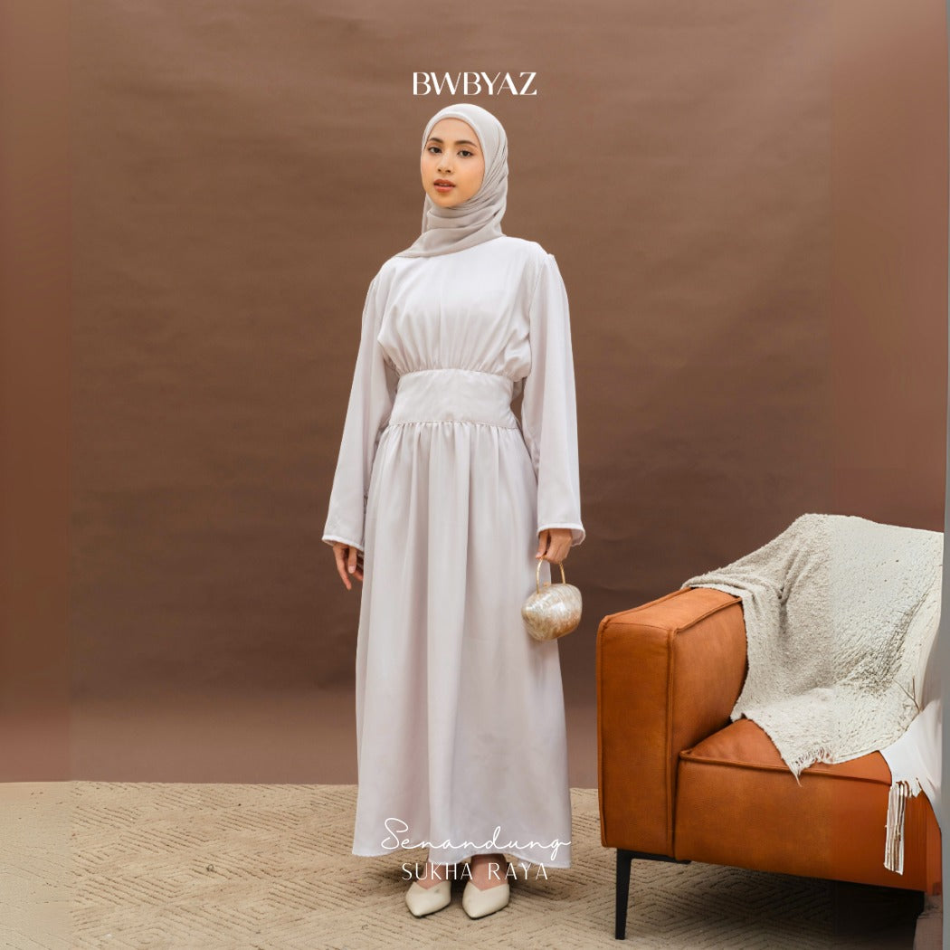 A woman wearing a beige Anisha Obi dress from BWBYAZ with a hijab, standing next to a brown chair with a beige dress on the backrest.