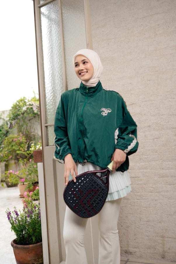 Woman in green windbreaker jacket by Haidee Orlin and hijab holding a black bag, standing indoors with plants outside.