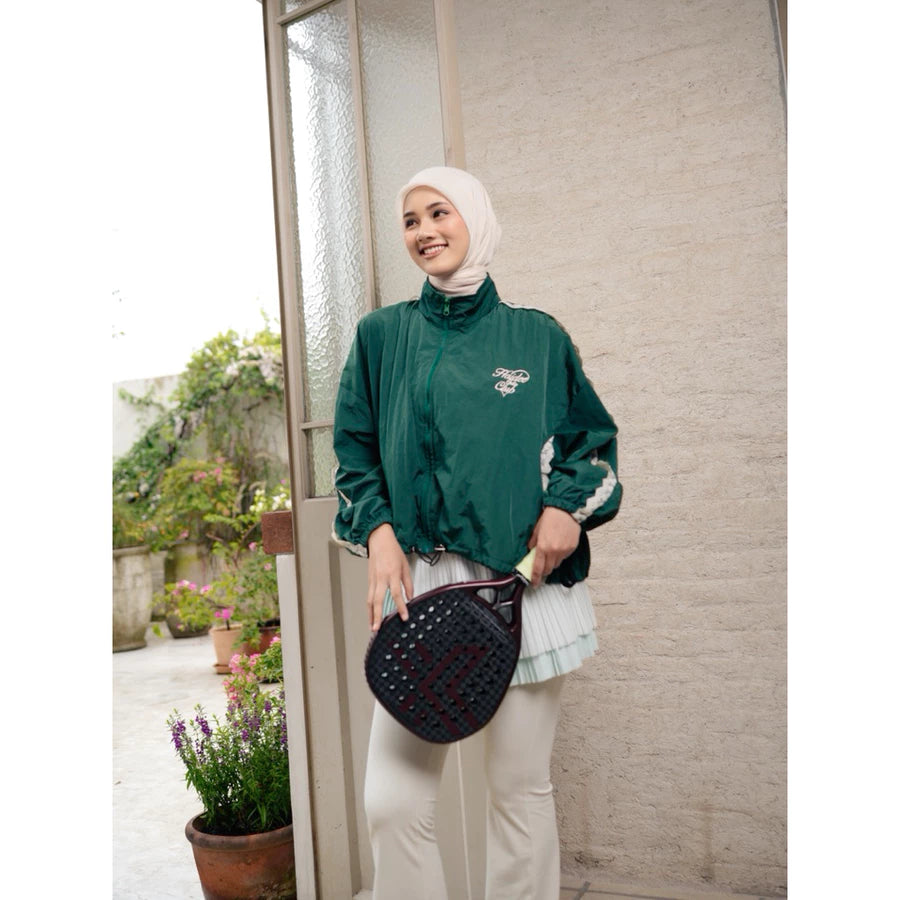 Woman in green windbreaker jacket by Haidee Orlin and hijab holding a black bag, standing indoors with plants outside.