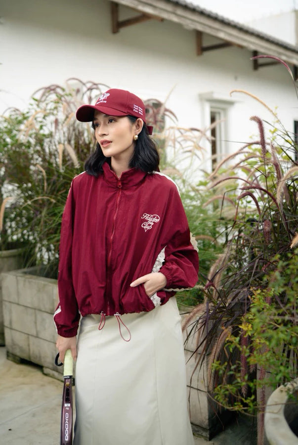Person wearing a maroon windbreaker jacket by Haidee Orlin and cap holding a tennis racket in an outdoor setting