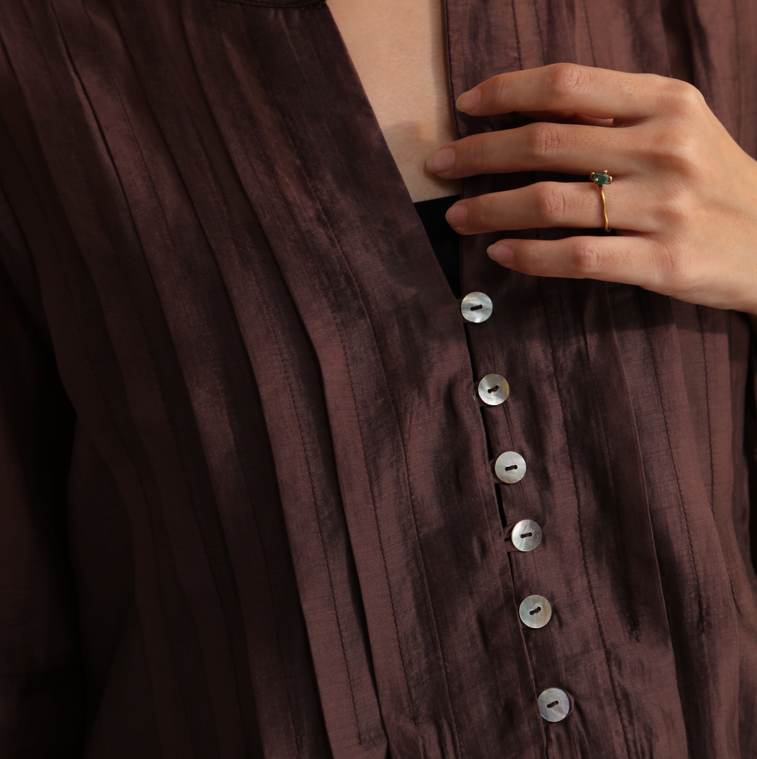 Close-up of a person wearing a dark brown blouse by Kasyalia with buttons and a ring on a dark background
