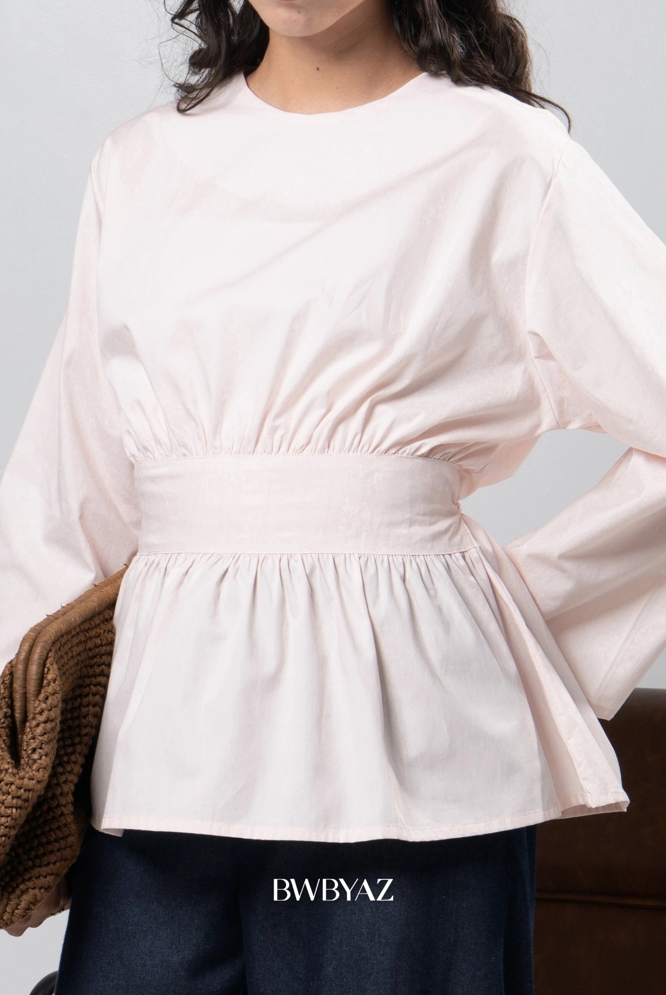 Person wearing a light pink blouse with long sleeves by BWBYAZ on a gray background, holding a brown bag.