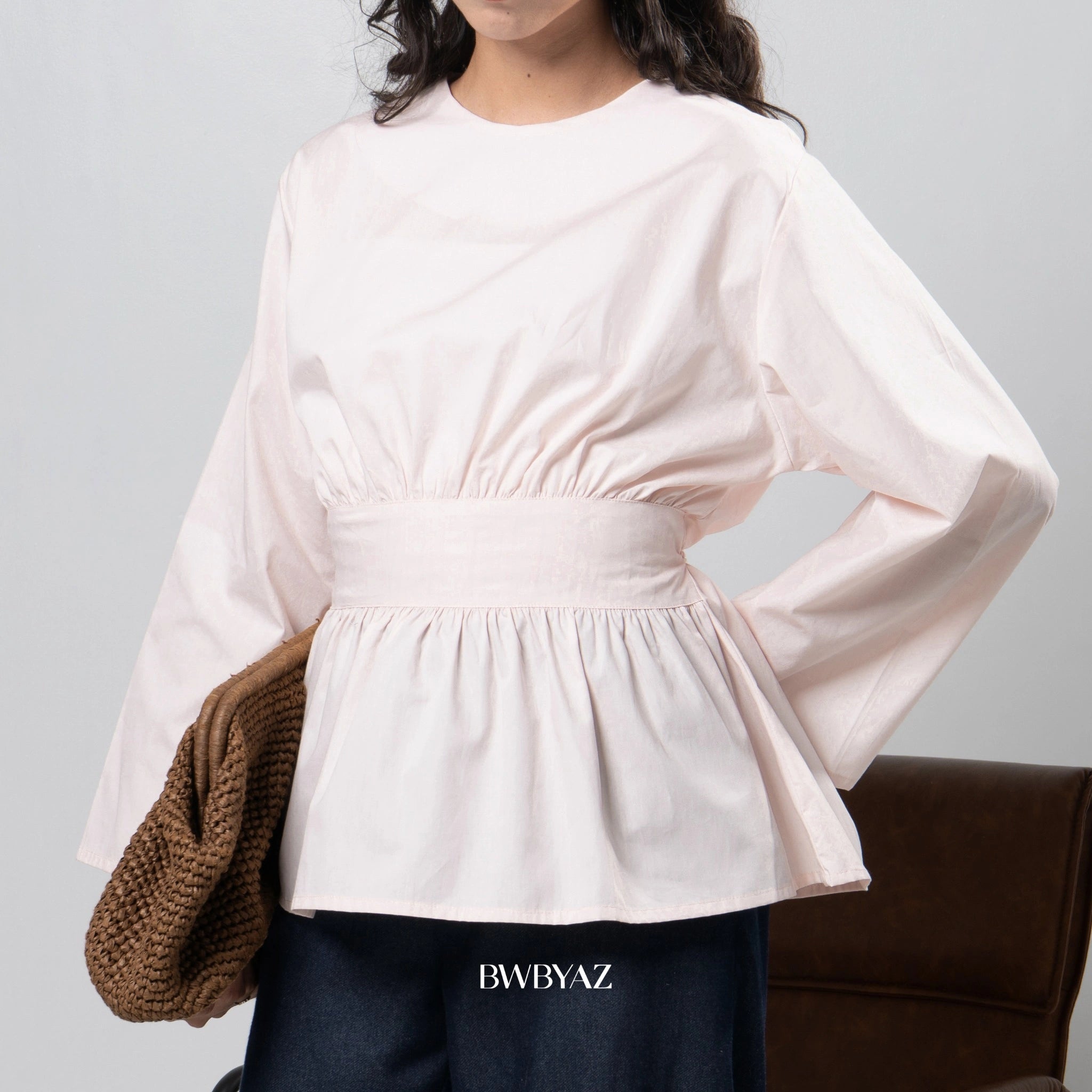 Person wearing a light pink blouse with long sleeves by BWBYAZ on a gray background, holding a brown bag.