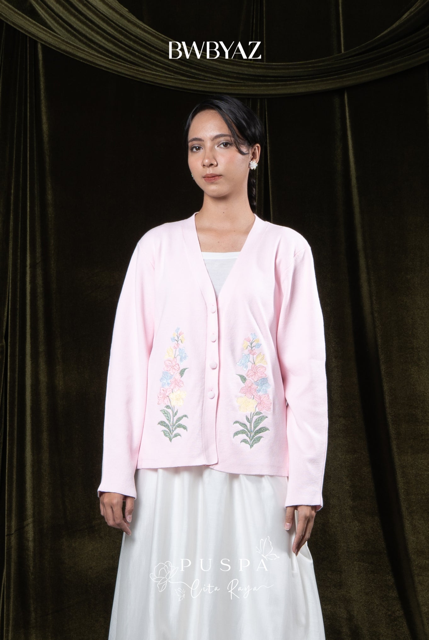 Woman wearing a pink embroidered cardigan over a white outfit against a dark curtain background with 'BWBYAZ' branding.