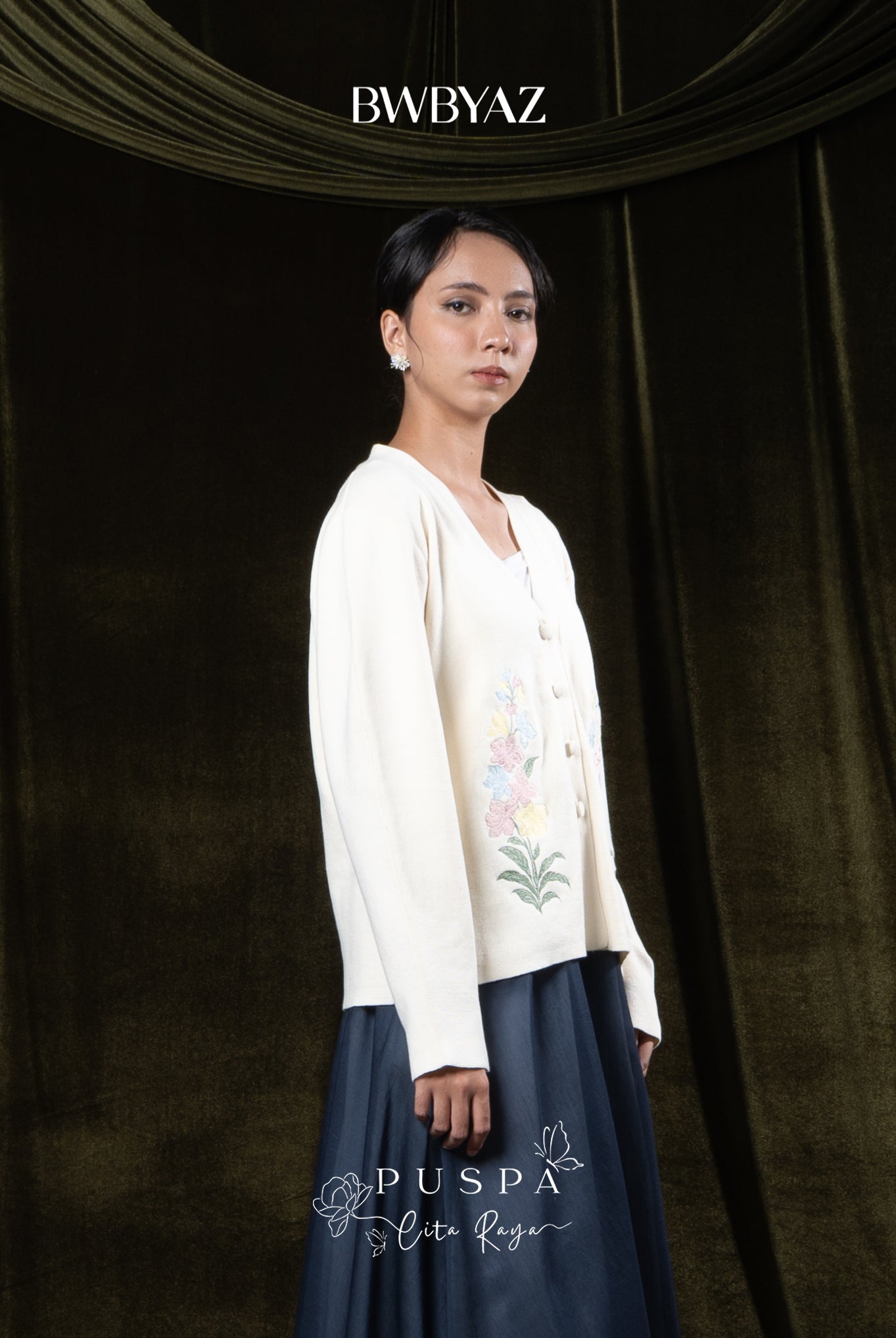 Woman wearing a cream-colored embroidered cardigan over a white outfit against a dark curtain background with 'BWBYAZ' branding.