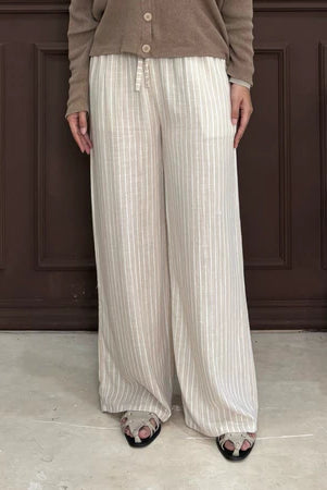 Person wearing a brown cardigan and nude pants with white stripe by Renaette standing against a dark paneled wall.