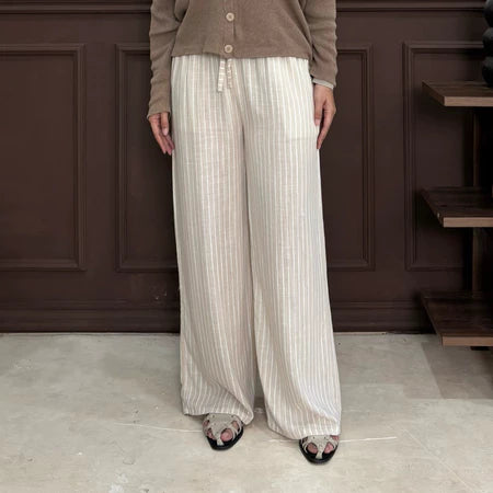 Person wearing a brown cardigan and nude pants with white stripe by Renaette standing against a dark paneled wall.