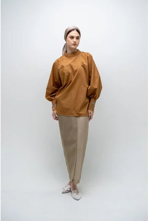 A person wearing a brown, long-sleeved blouse by Glashka with a round neckline and decorative buttons on the cuffs. The blouse has a loose fit and the person is paired with black pants.