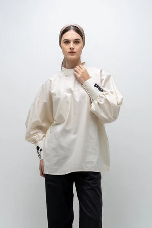 A person wearing a white, long-sleeved blouse by Glashka with a round neckline and decorative buttons on the cuffs. The blouse has a loose fit and the person is paired with black pants.