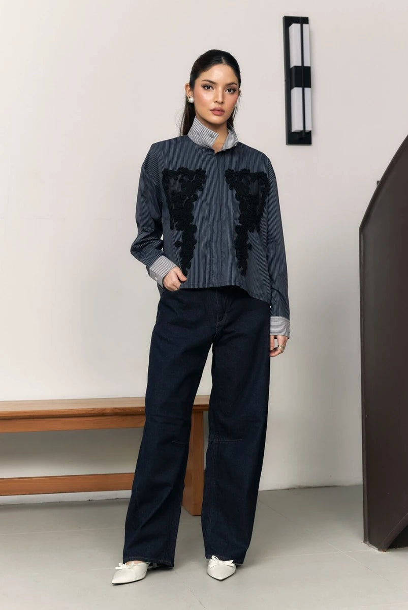 Woman wearing a navy shirt with black floral embroidery by Studio Tui and dark pants in a minimal setting.