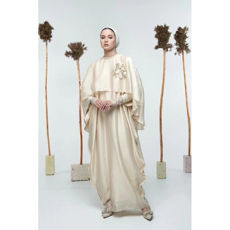 A person wearing a beige maxi dress by Glashka with long sleeves standing against a white background with decorative trees in the background.