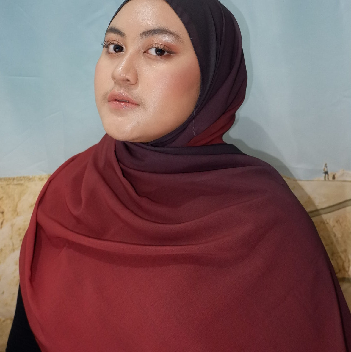 Person wearing a maroon pashmina hijab and top against a light blue background
