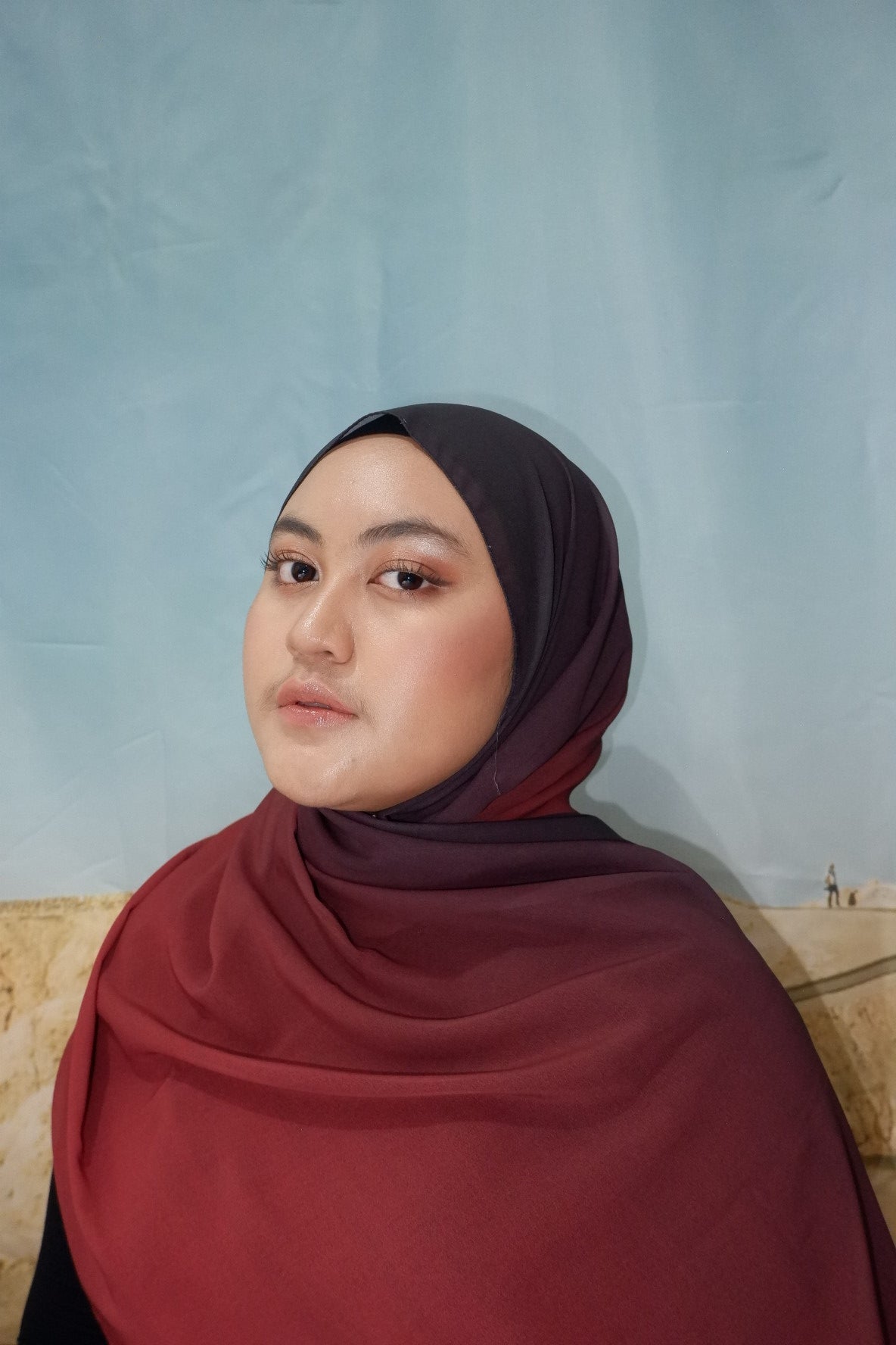 Person wearing a maroon pashmina hijab and top against a light blue background