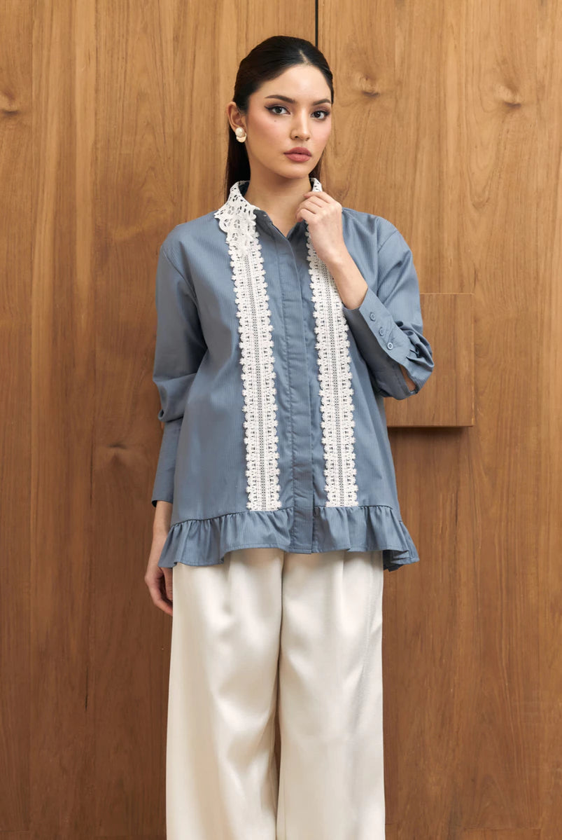 A woman models a dusty blue long-sleeve shirt by Studio Tui with white embroidered vertical panels down the front, paired with cream wide-leg trousers, standing against a wooden wall.