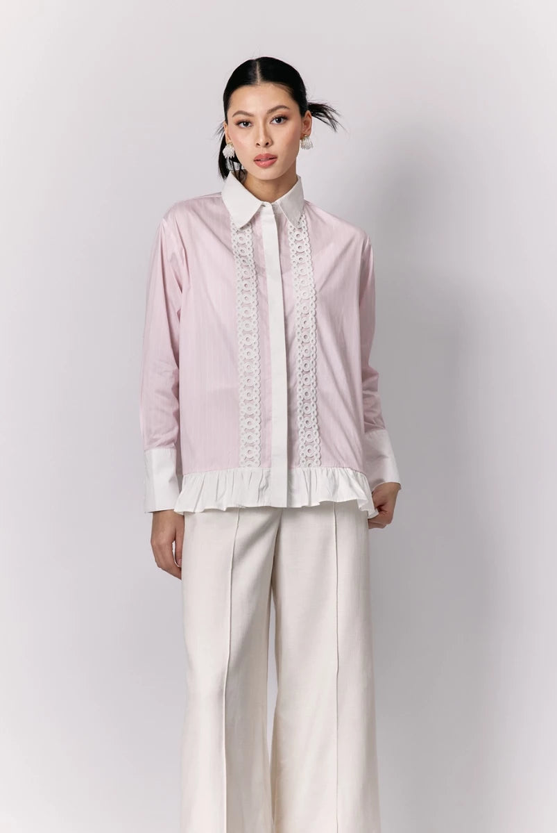 Woman wearing a light pink shirt by Studio Tui with lace details and white pants on a plain background