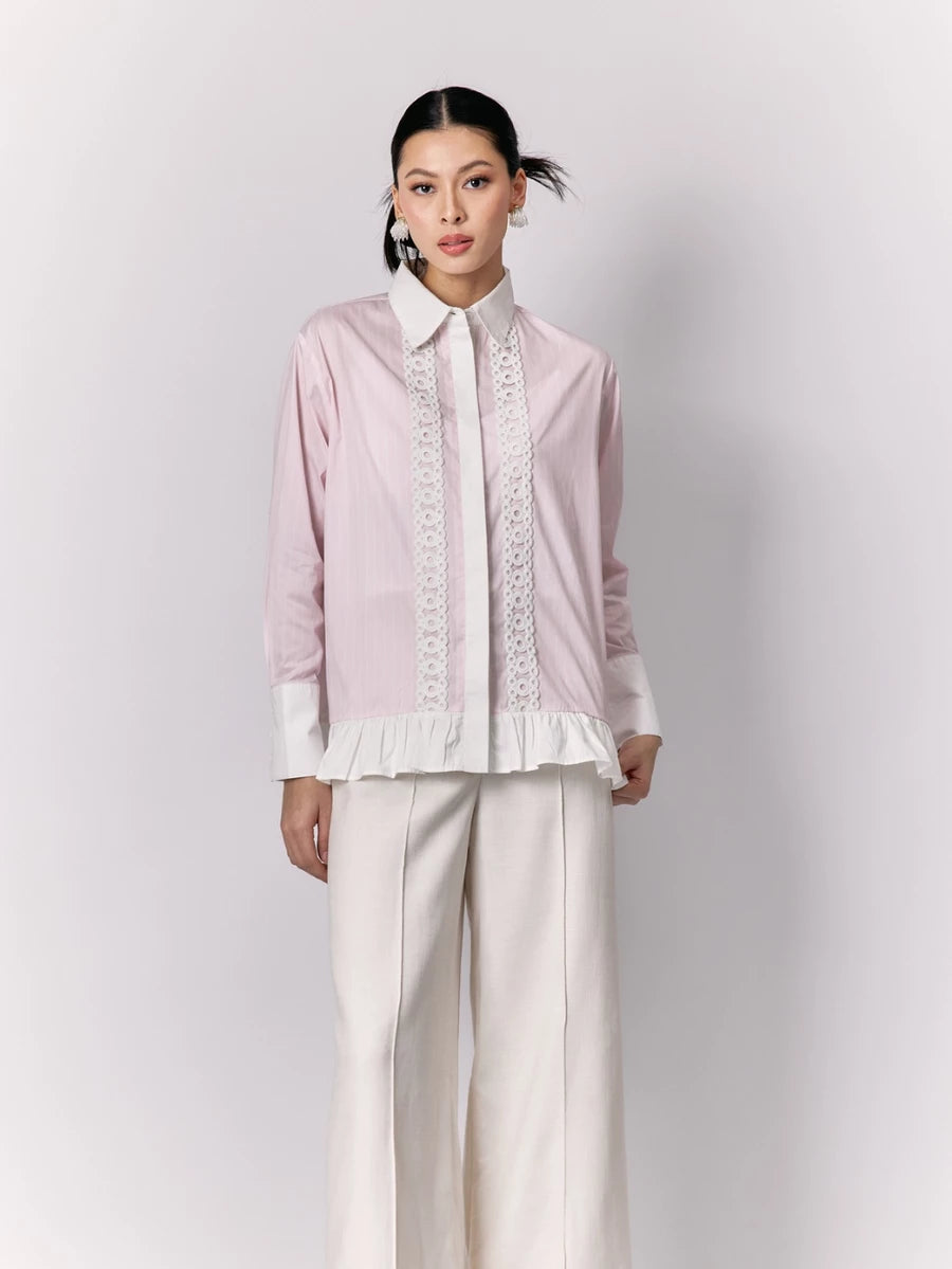 Woman wearing a light pink shirt by Studio Tui with lace details and white pants on a plain background
