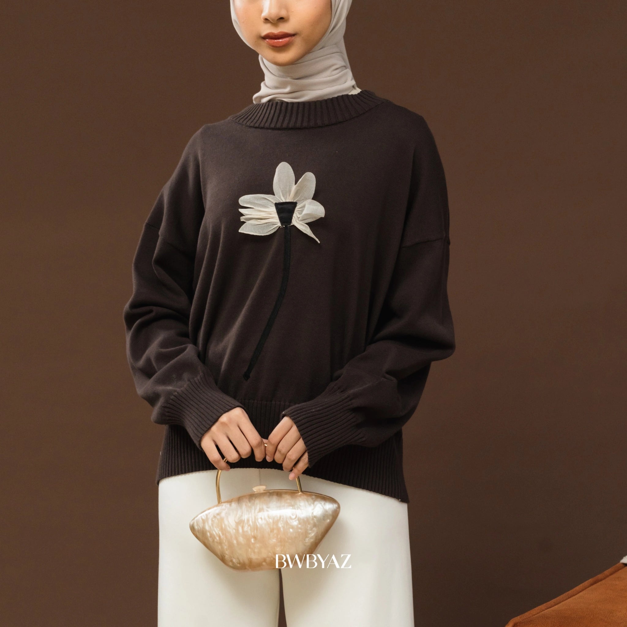 Person wearing a dark brown sweater with a floral design from BWBYAZ, holding a beige handbag against a neutral background.