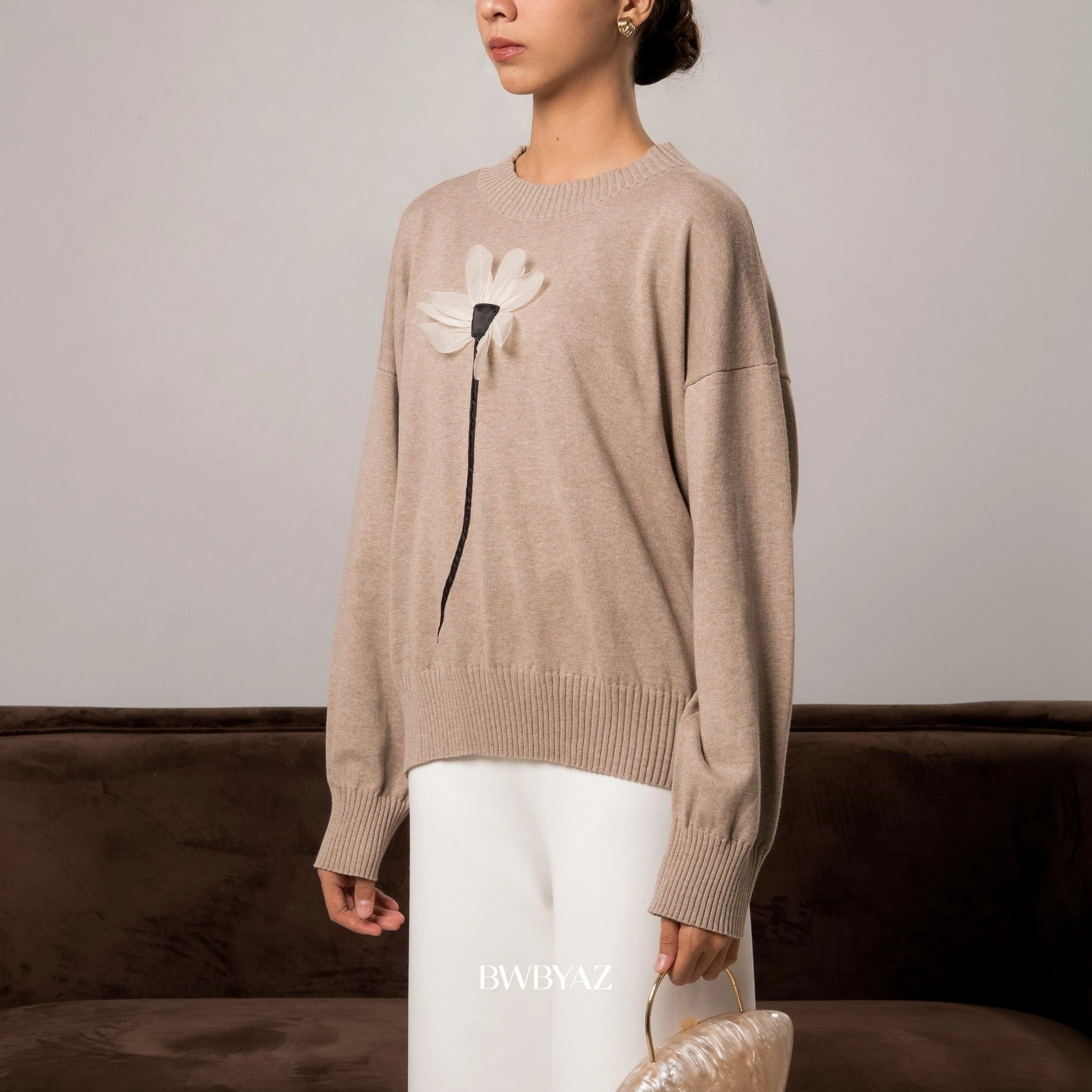 Person wearing a light brown sweater with a floral design from BWBYAZ, holding a beige handbag against a neutral background.