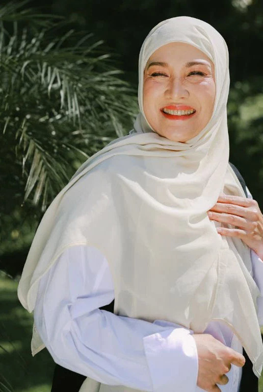 A woman wearing a light yellow pashmina headscarf over a black outfit against a grey background.