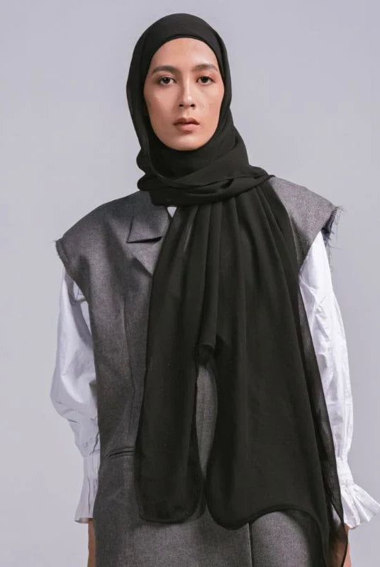 A woman wearing a black pashmina headscarf over a black outfit against a grey background.