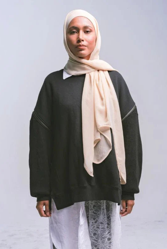 A woman wearing a beige pashmina headscarf over a black outfit against a grey background.