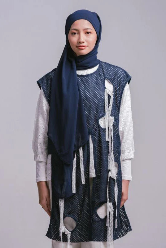 A woman wearing a dark blue pashmina headscarf over a black outfit against a grey background.