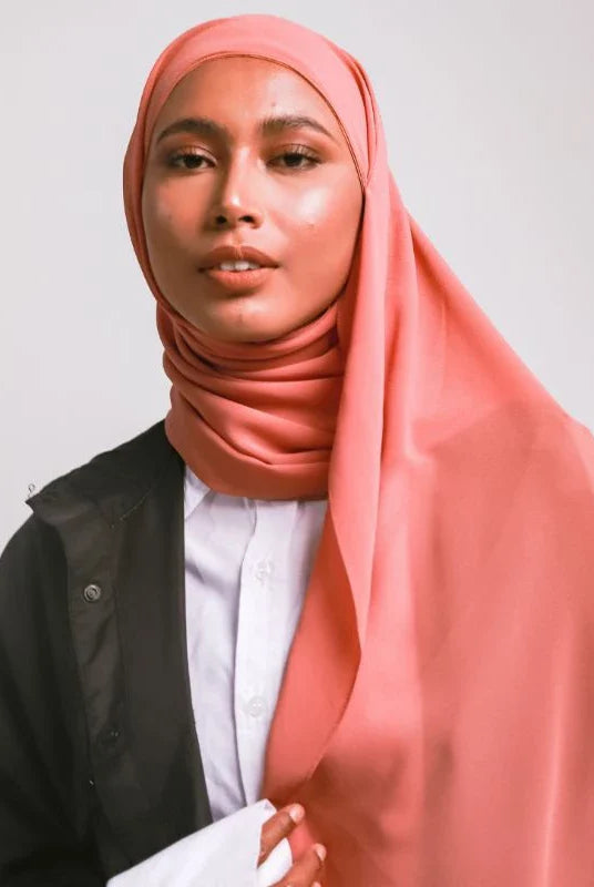 A woman wearing a taupe-colored pashmina headscarf over a black outfit against a grey background.