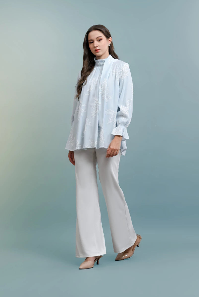 Woman wearing a light blue blouse with floral patterns by Kasa Heritage and white pants against a gray background