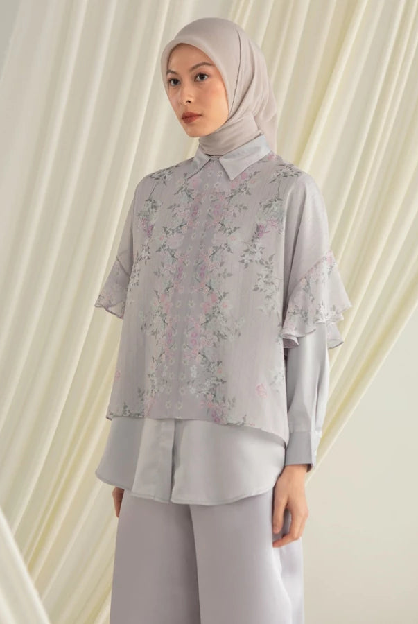 Woman wearing a light gray floral shirt by Hijabchic and pants with a matching hijab against a white curtain background.